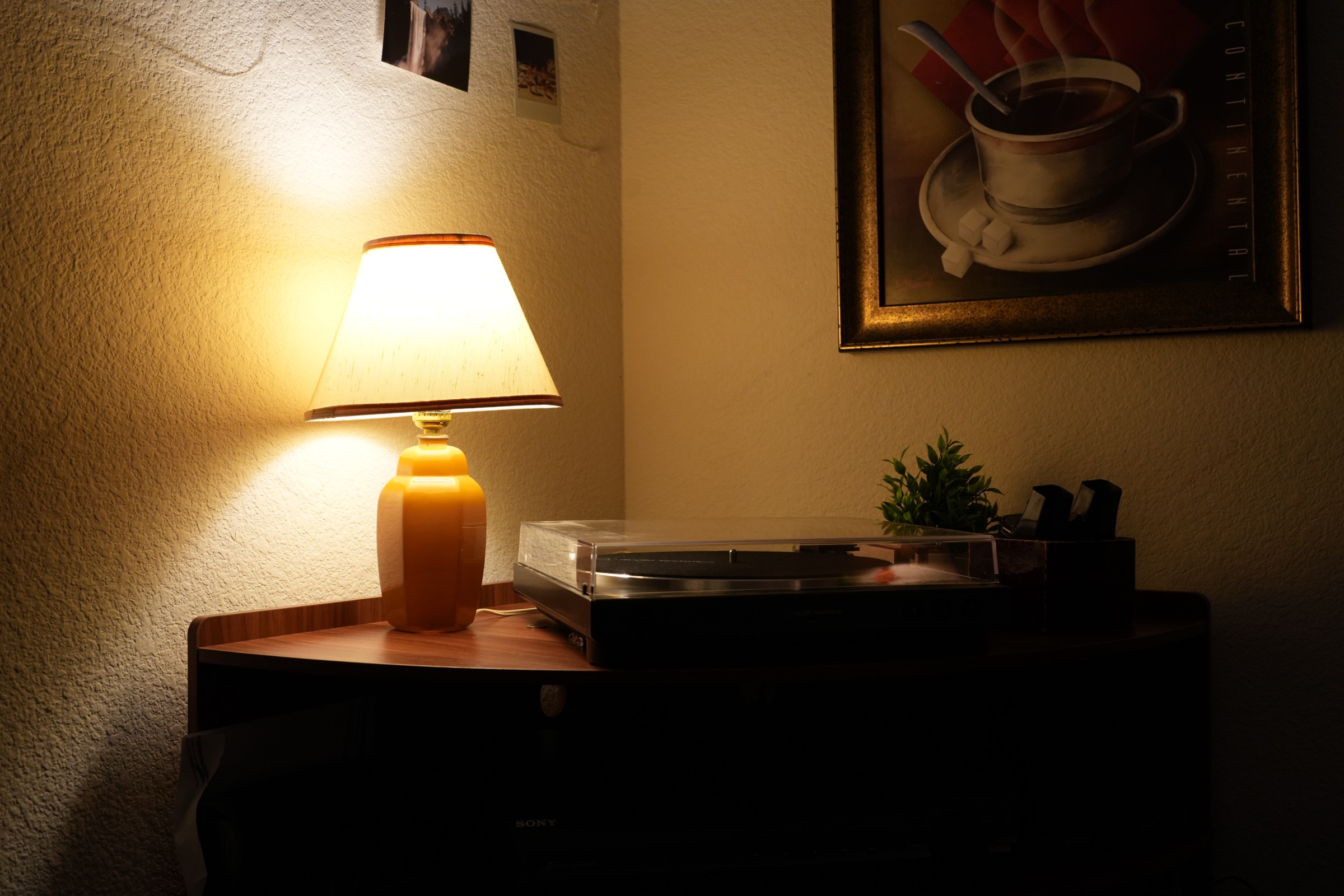 A warmly lit lamp beside a record player, with framed coffee artwork hanging above it.