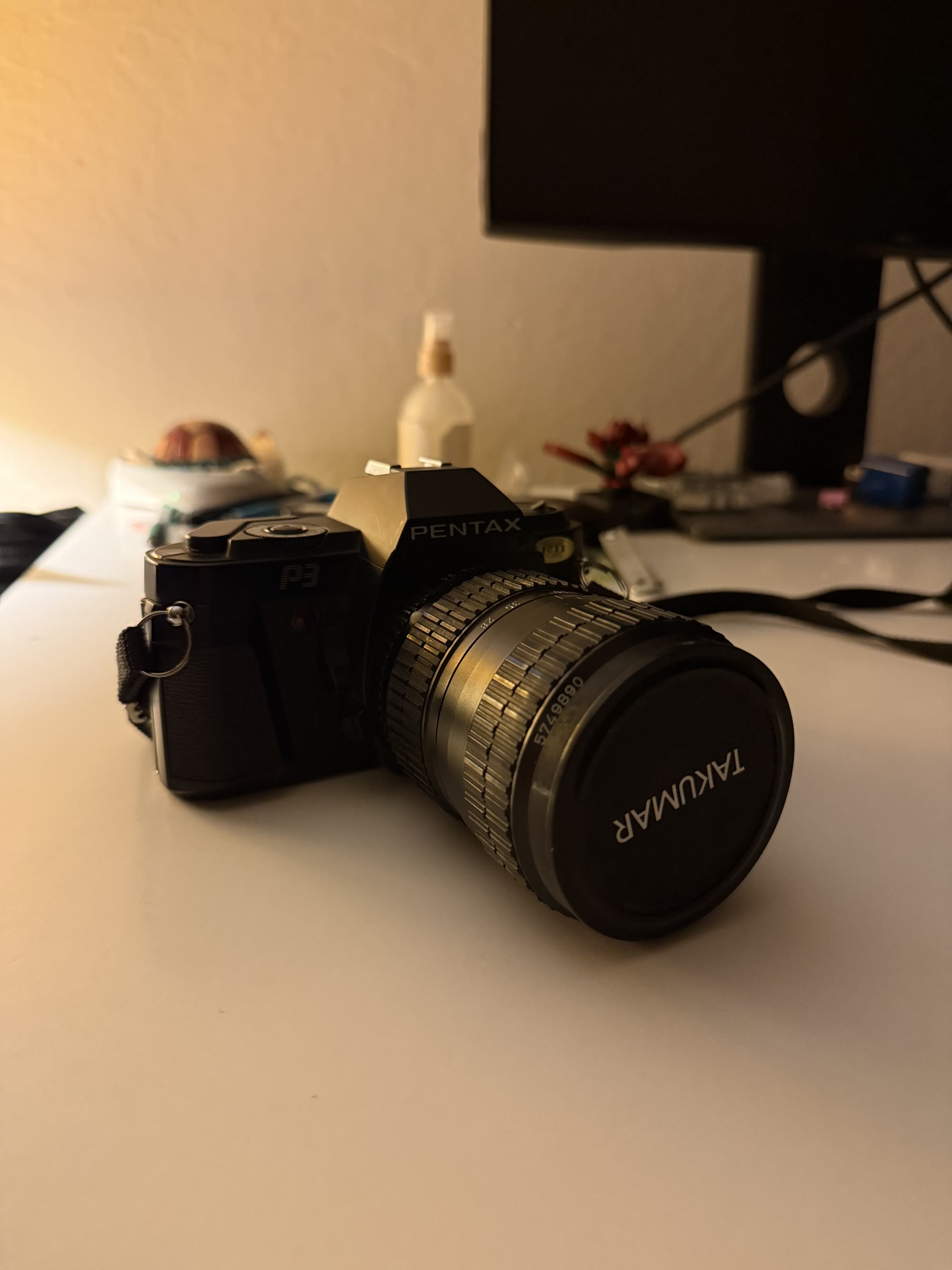 A black Pentax P3 film camera with a long lens sitting on a desk in warm light.